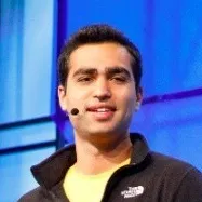 Man with short dark hair wearing a black jacket and yellow shirt speaking with a microphone headset against a blue background.