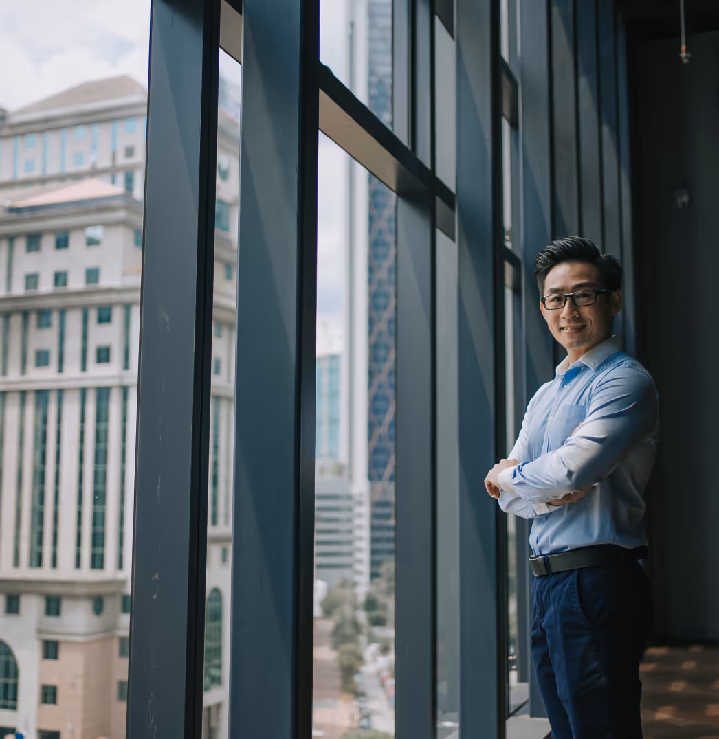 Smiling man in glasses and blue shirt stands with arms crossed by tall office windows.