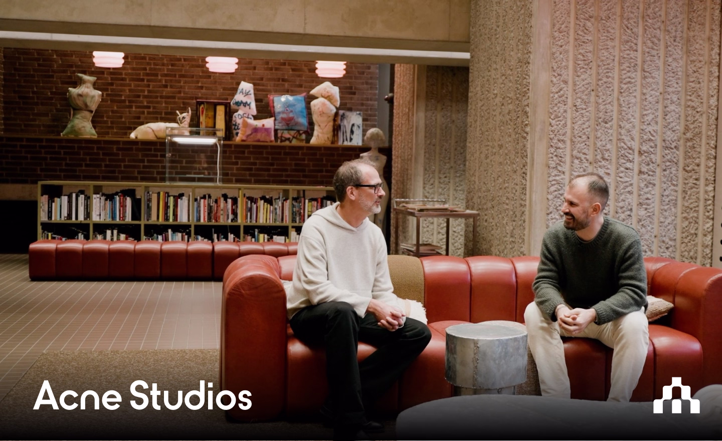 Two men talking on a red sofa, with the Acne Studios logo overlay