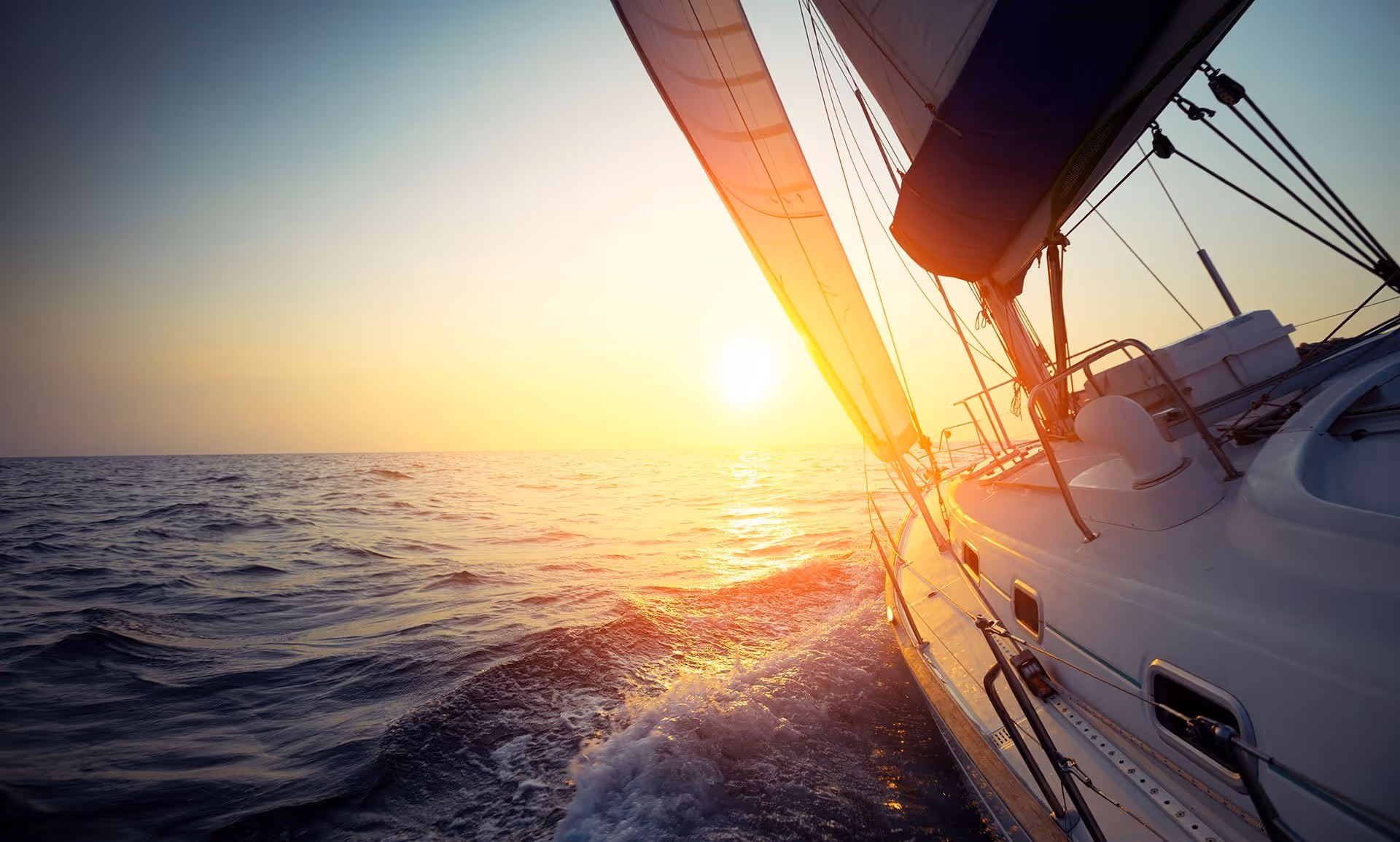 A sailboat moves through the ocean at sunset, with the sun casting a warm glow.