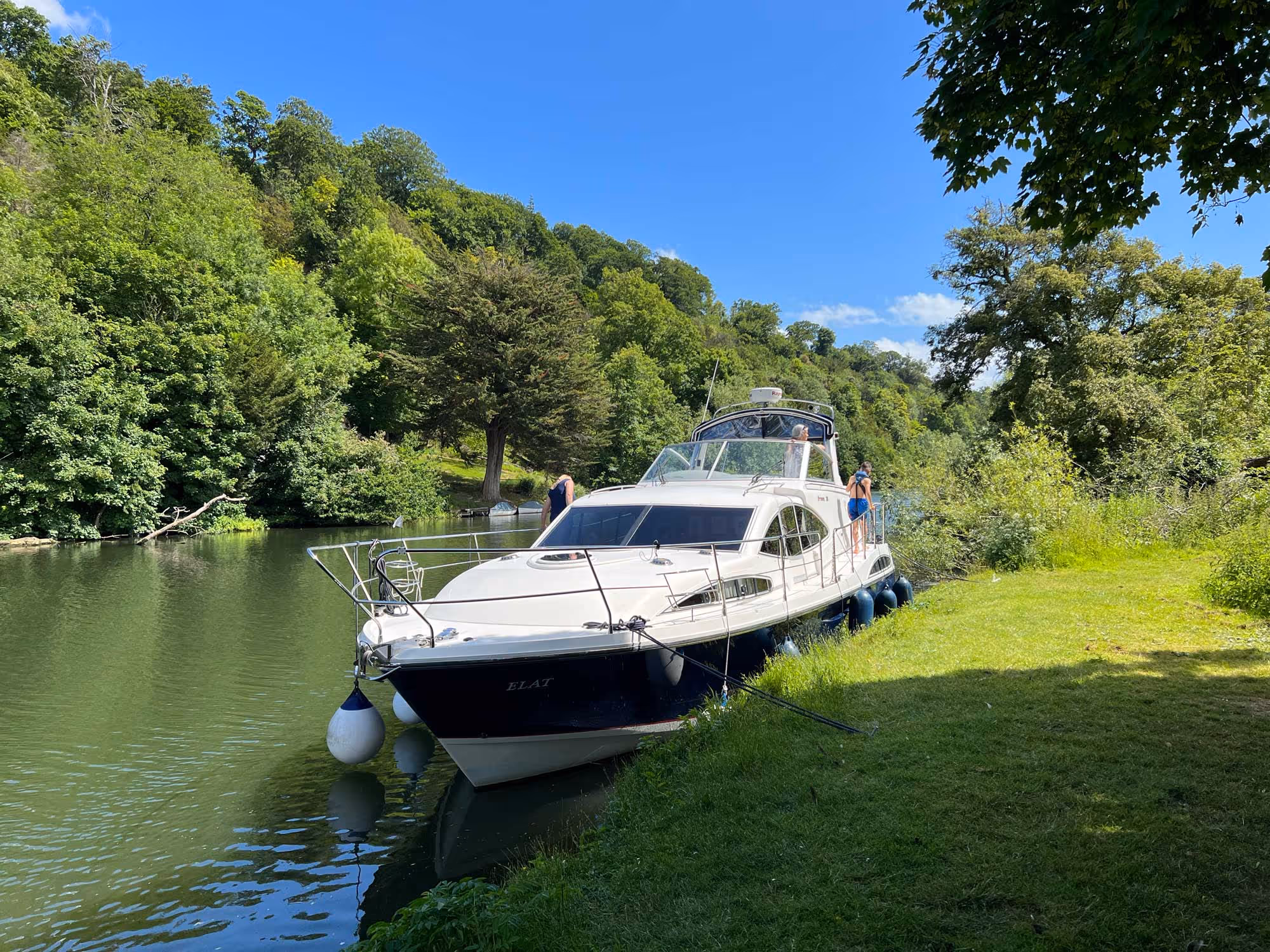 A white boat is docked along a riverbank with two people standing nearby.