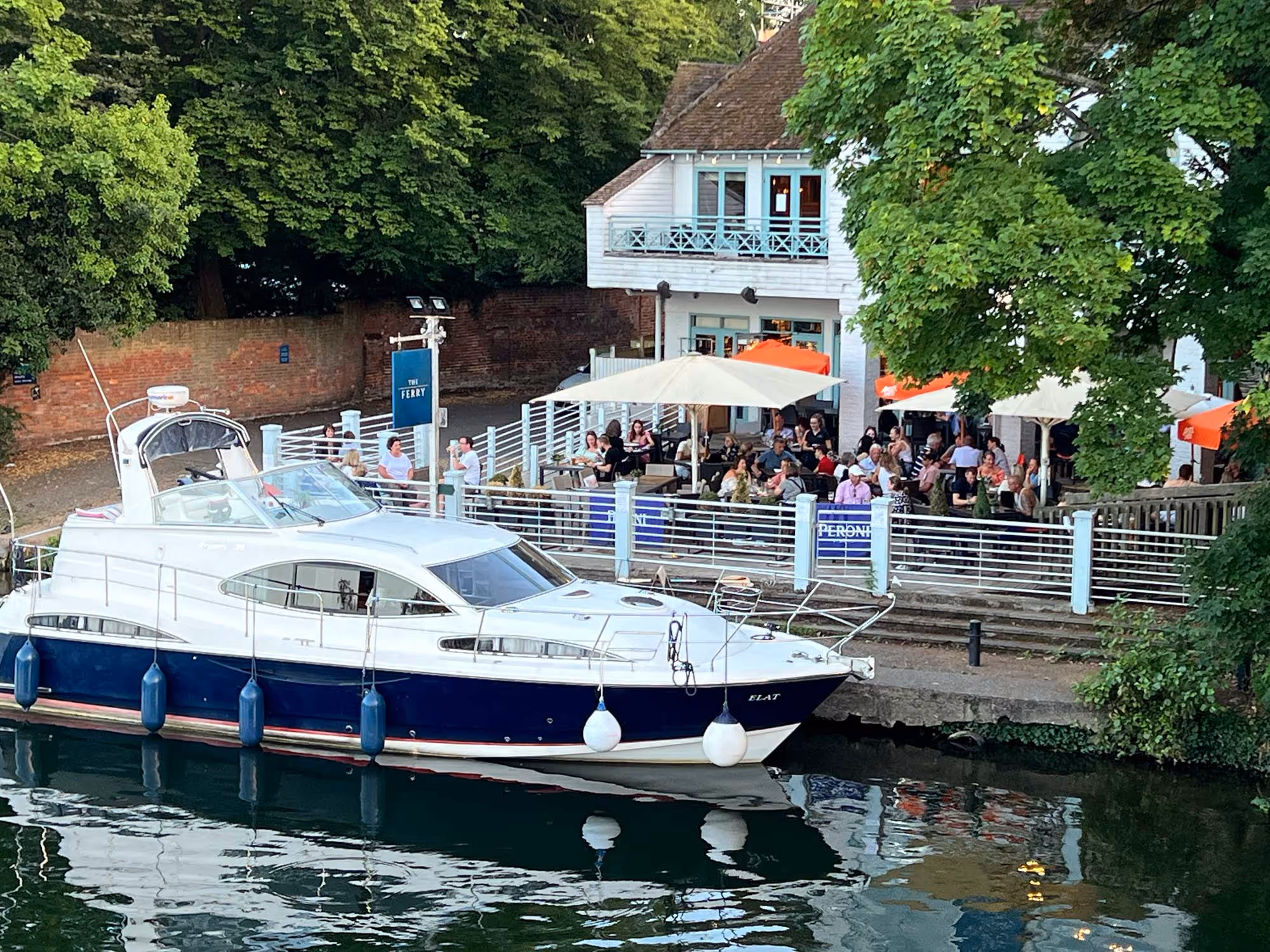 A boat docked near a riverside restaurant with people dining outdoors under umbrellas.