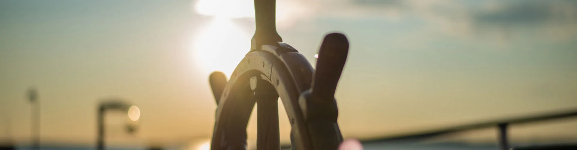 Close-up of hands gripping a ship's wheel against a bright sunset sky.