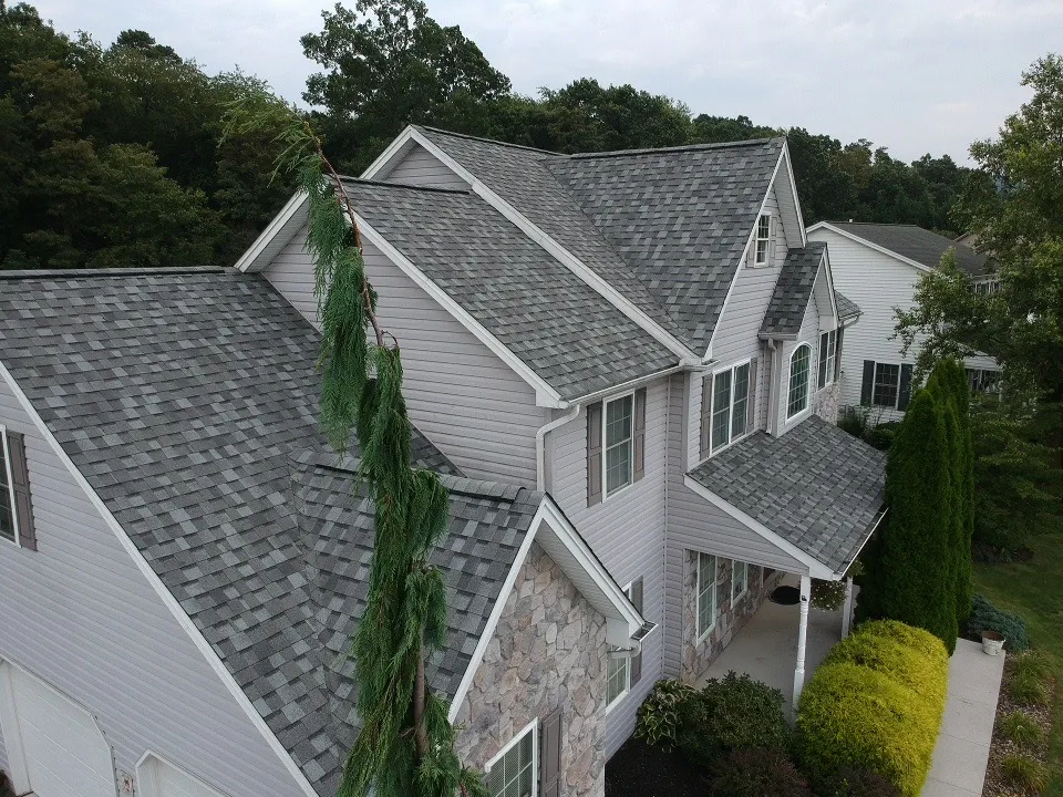 Aerial view of a two-story suburban home with gray asphalt shingle roofing, light purple siding, and stone exterior accents, surrounded by trees and neatly trimmed landscaping.