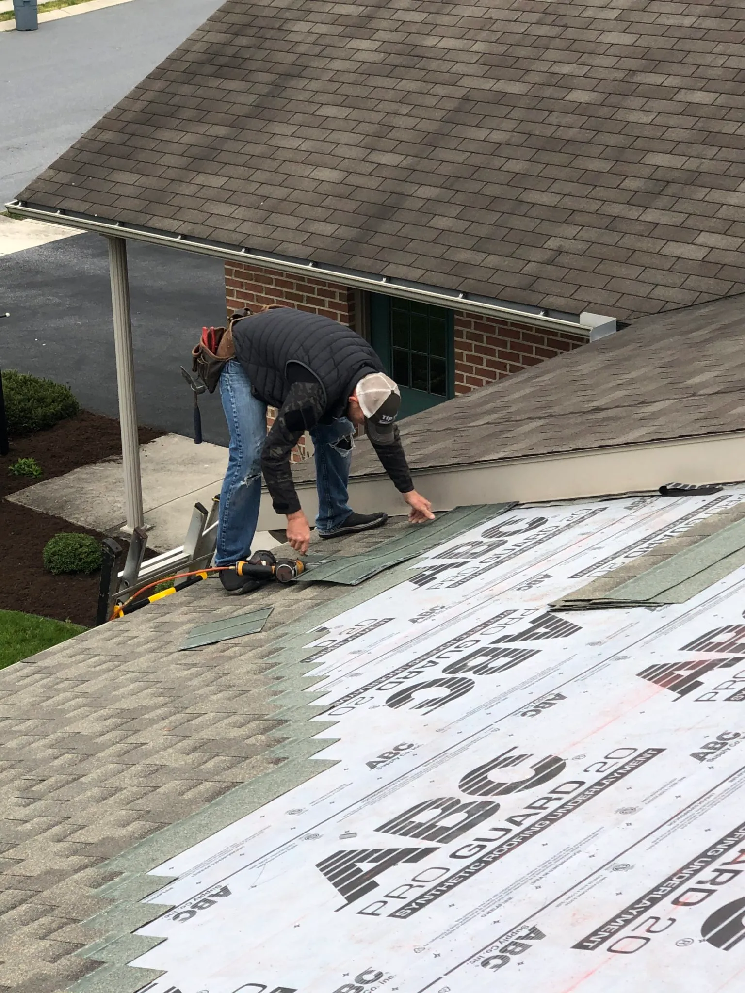 Roofer installing asphalt shingles over ABC Pro Guard 2.0 roofing underlayment on a sloped residential roof, using a nail gun and wearing safety gear and a tool belt.