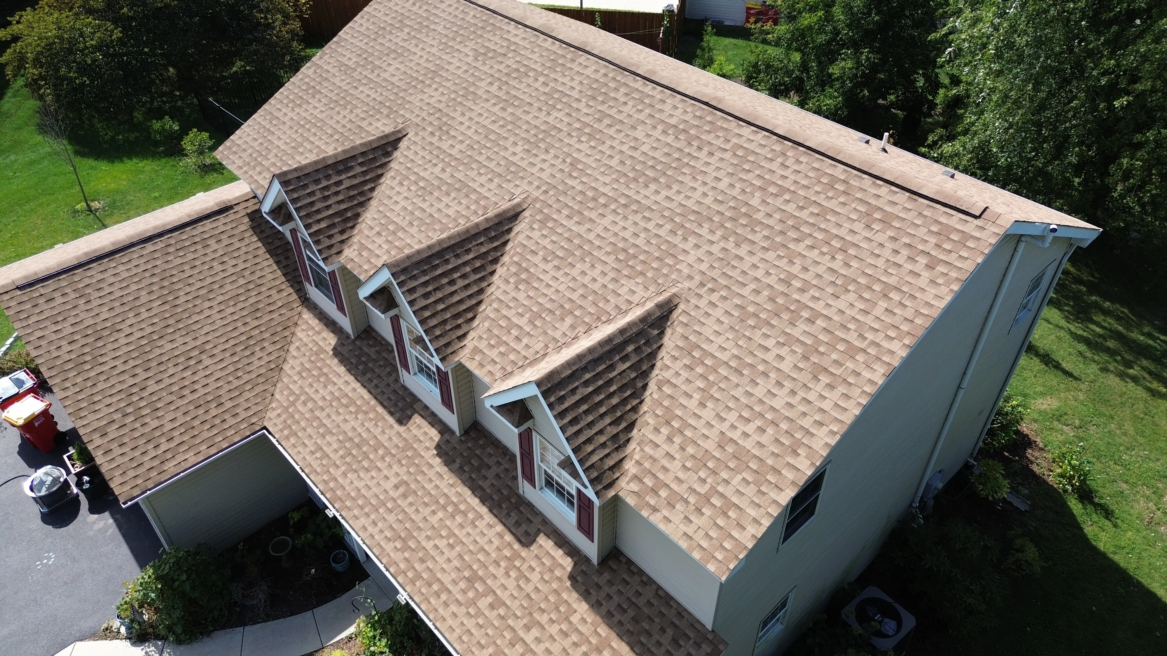 Aerial view of a residential home featuring a freshly installed light brown asphalt shingle roof with three gabled dormers and red window shutters, surrounded by green trees and lawn.
