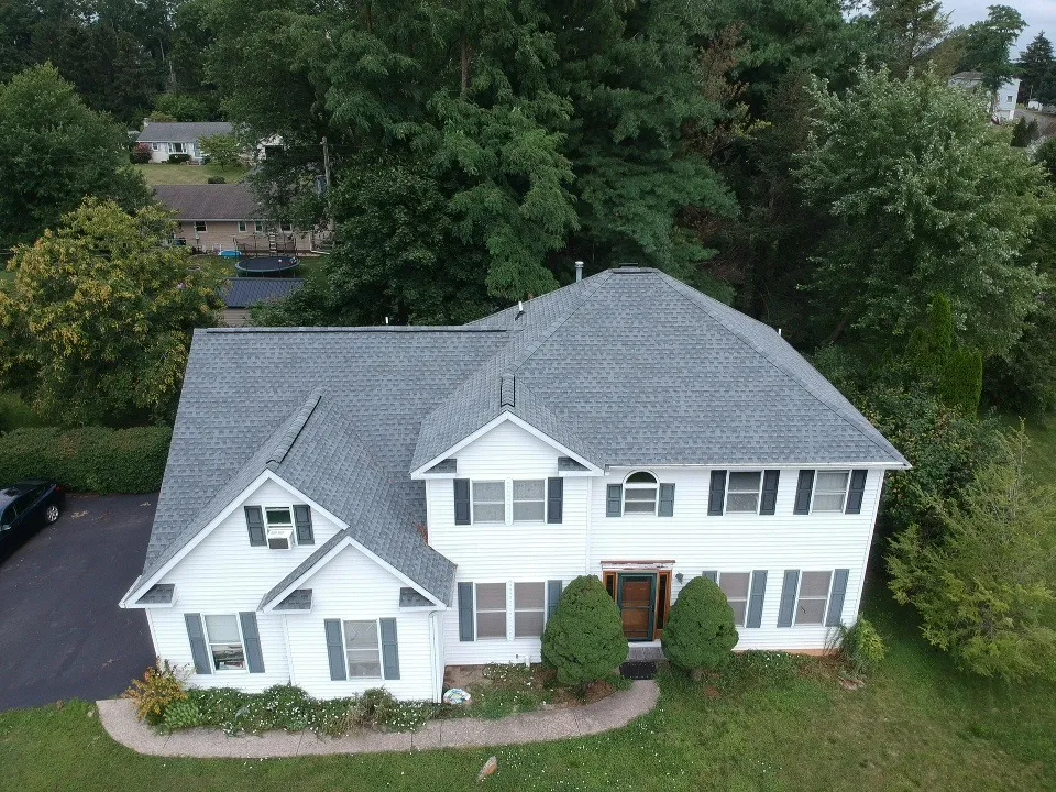 Aerial view of a two-story white suburban house with a dark gray asphalt shingle roof, black shutters, and a landscaped front yard bordered by trees and a paved driveway.