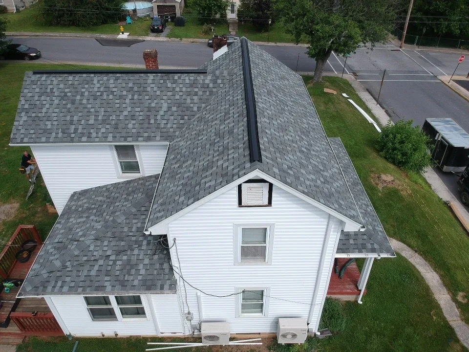 Top-down view of a white two-story house with a newly installed dark gray asphalt shingle roof, surrounded by a grassy yard and adjacent to a quiet residential street with visible traffic lines and sidewalks.