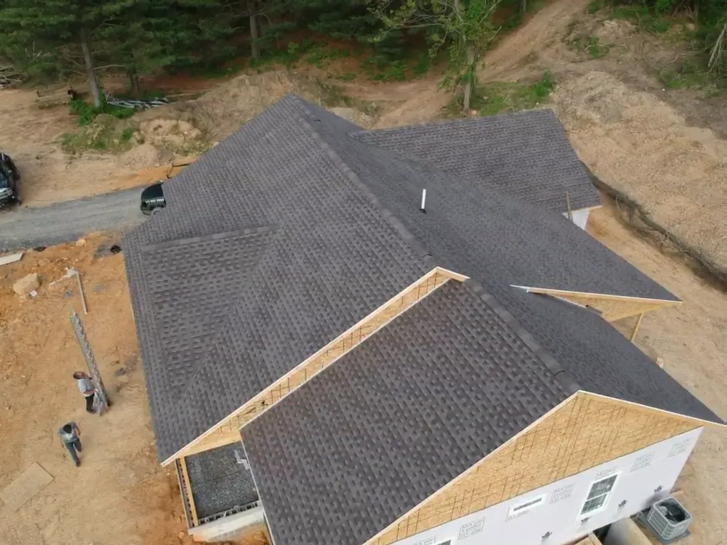 Aerial view of a newly constructed home with dark asphalt shingles installed on a complex roof structure, surrounded by construction materials and dirt terrain.