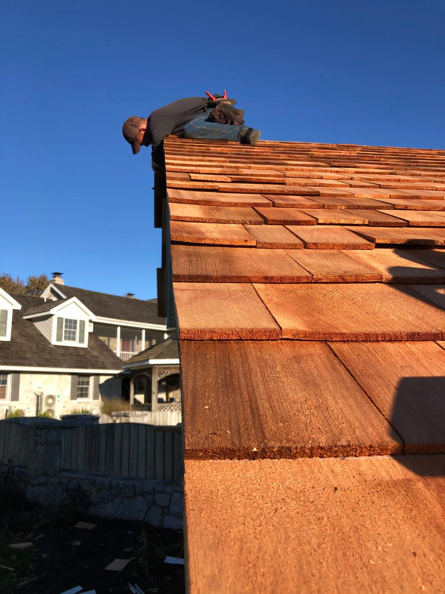 Roofer installing wooden shingles on a steep rooftop in clear weather, with a large residential house and blue sky in the background.