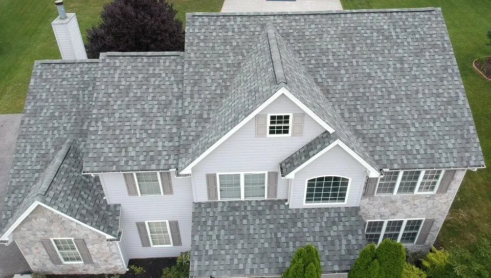 Aerial view of a two-story home with gray asphalt shingles, multiple gables and dormers a stone facade, and front lawn below.