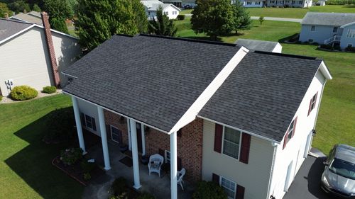 Aerial view of suburban home with dark asphalt shingles, columned porch, brick and siding exterior, and spacious lawn