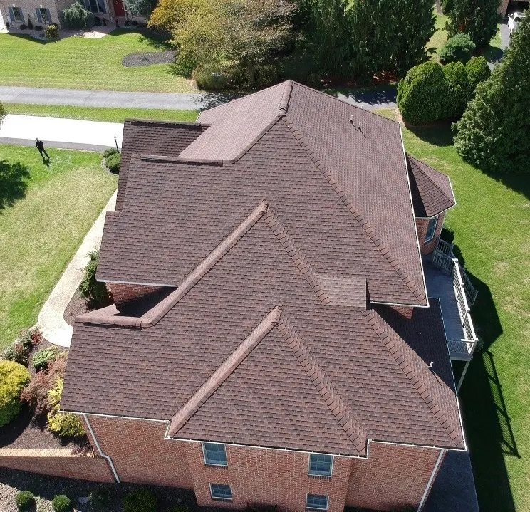 Aerial view of large brick home with brown shingle roof, complex gables and ridges, surrounded by a manicured lawn and trees.