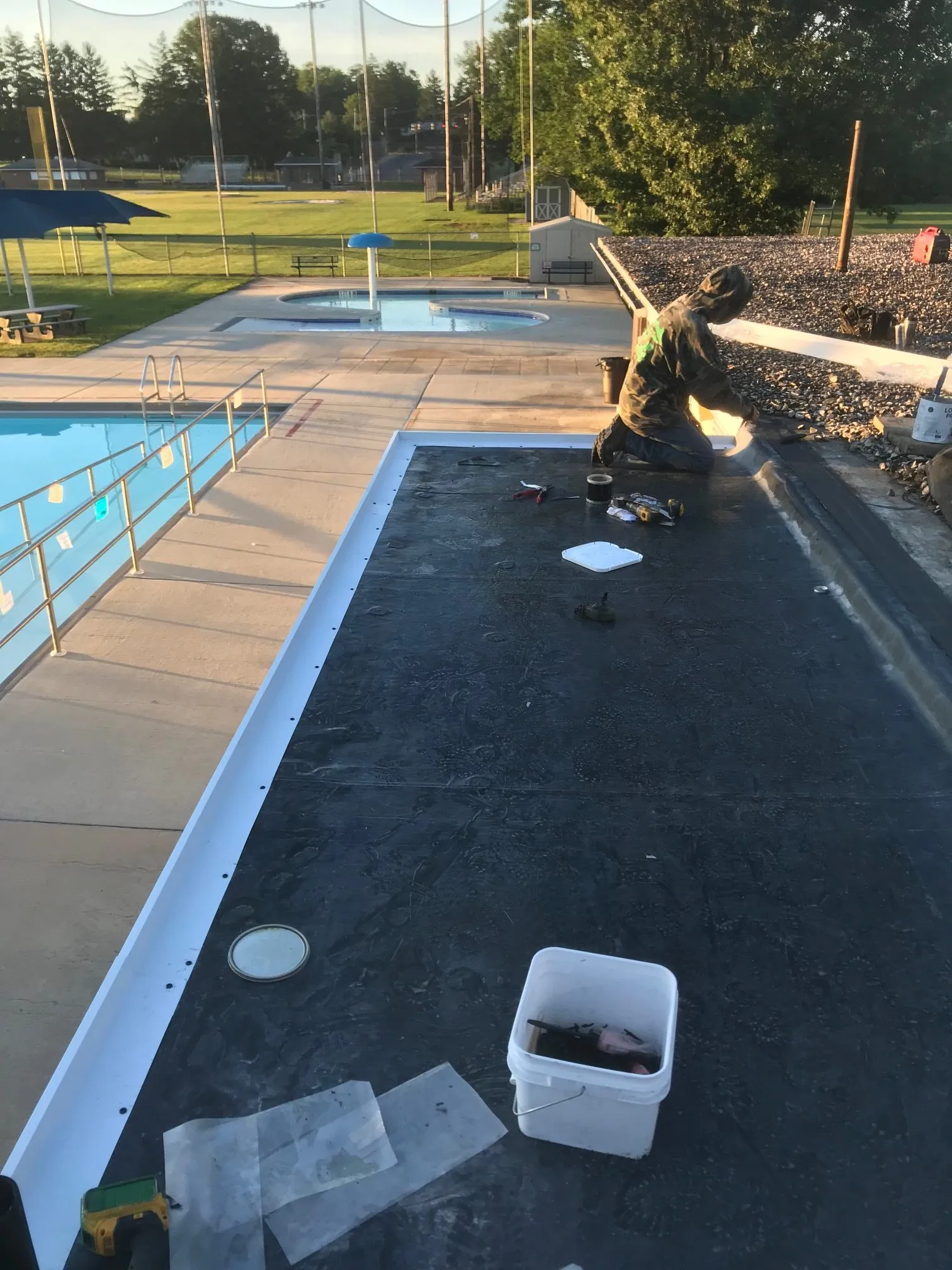 Technician installing EPDM rubber membrane on flat commercial roof beside outdoor pool, tools and bucket visible at dusk.