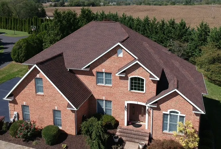 Aerial view of brick two-story home with brown GAF Hickory shingle roof, complex gables and ridges, landscaped yard, and trees in the background.