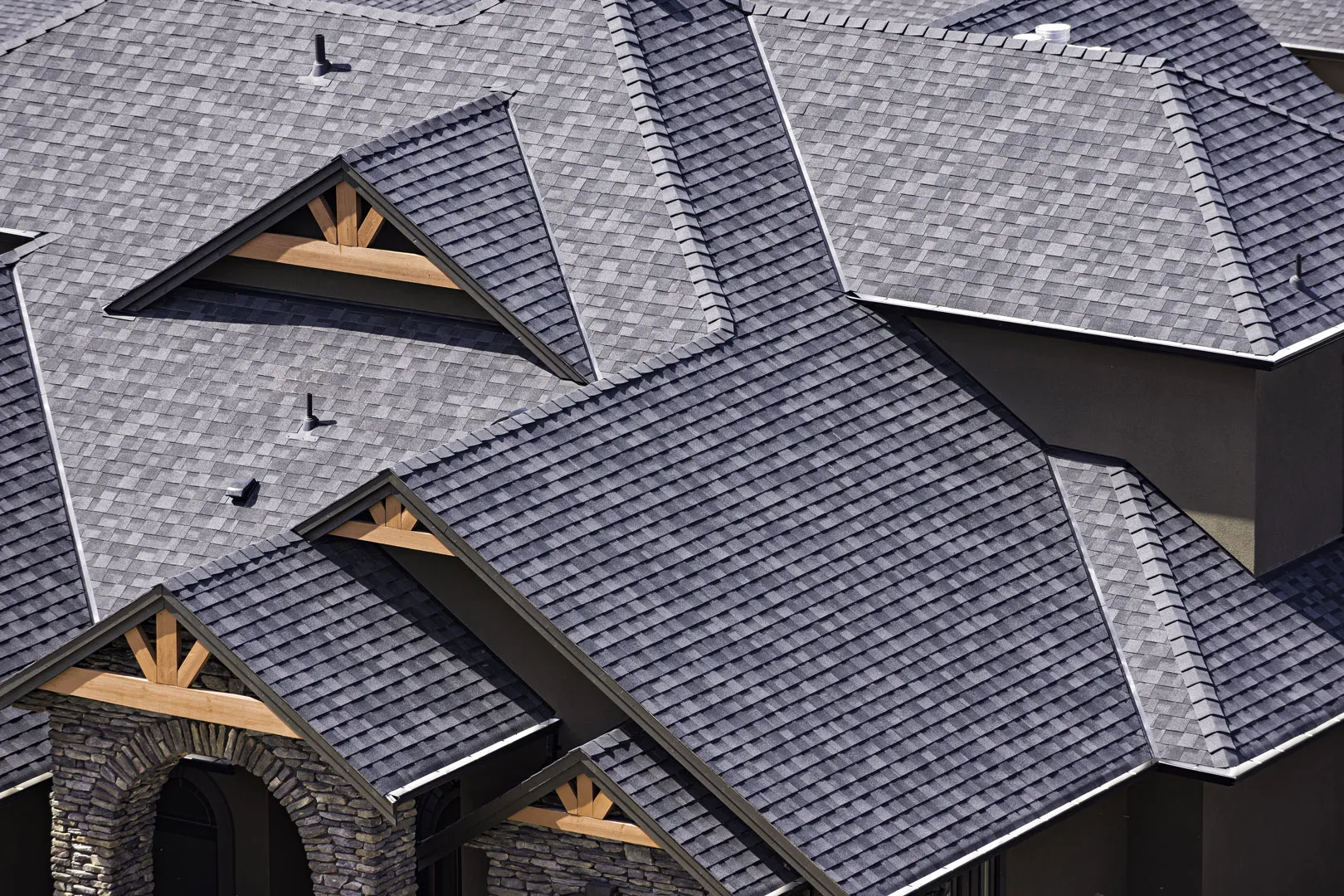 Close-up aerial view of complex asphalt shingle roof with many gables, valleys and timber accents on a luxury home.