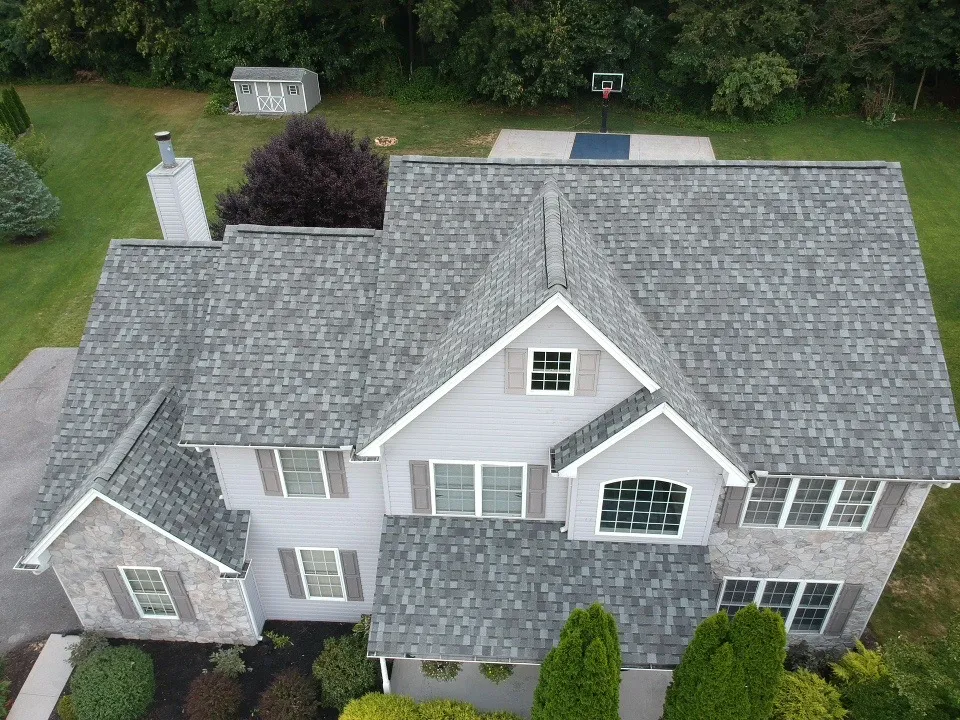 Aerial view of a two-story home with gray asphalt shingles, multiple gables and dormers a stone facade, and front lawn below.