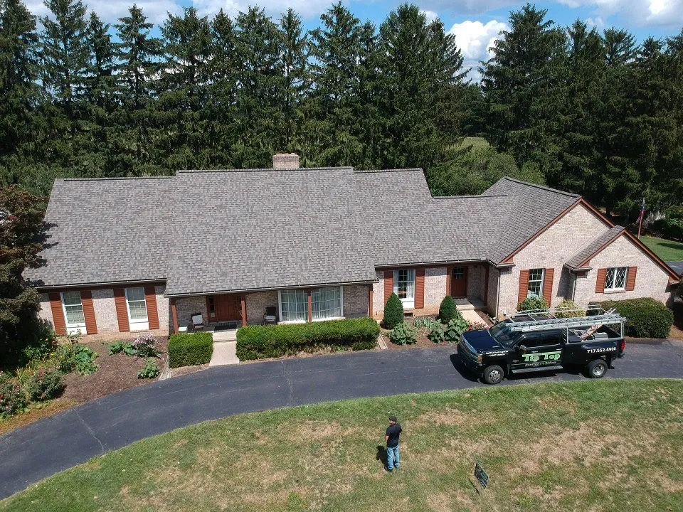 Aerial view of brick ranch-style home with new gray shingle roof; Tip Top Roofing service truck parked on curved driveway, tall pines behind.