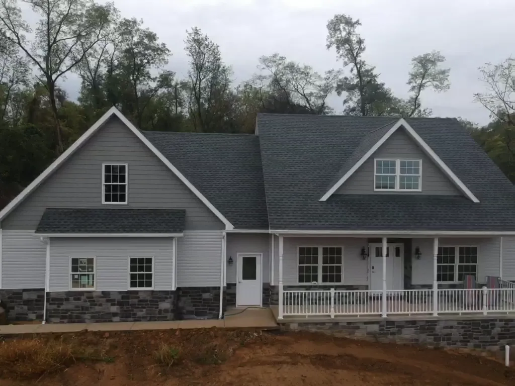 Front view of gray two-story home with dark shingle roof, white trim, stone veneer base, and wraparound porch, trees and cloudy sky behind.