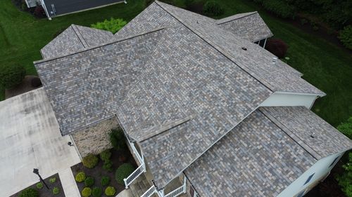 Aerial view of a large residential home with a newly installed multi-toned shingle roof, surrounded by green lawn, landscaping, and a concrete driveway.