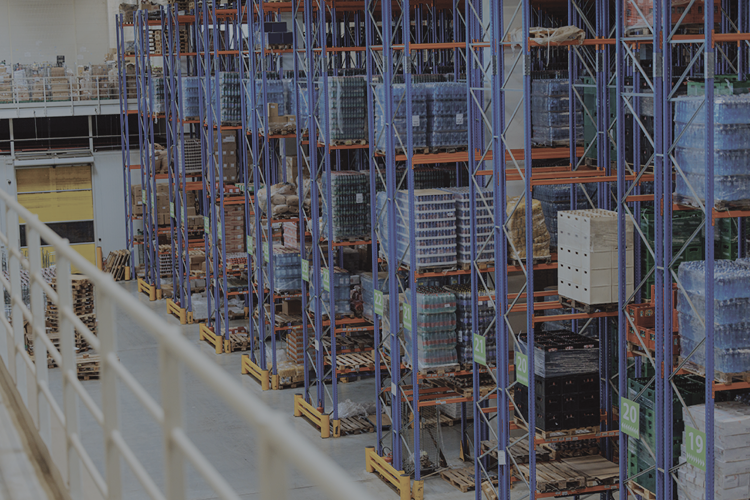 Warehouse aisle with tall industrial metal shelving racks storing pallets of bottled water and other goods.