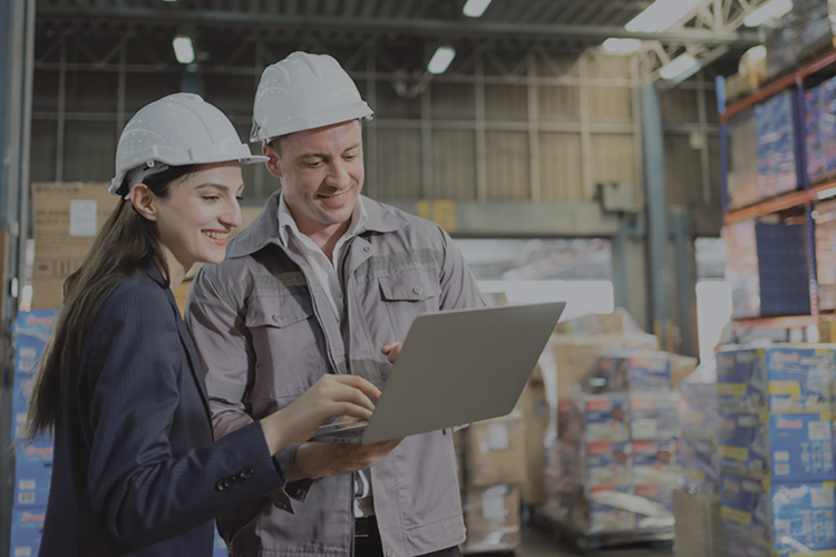 Two warehouse workers wearing hard hats looking at a laptop in a storage facility with stacked boxes.