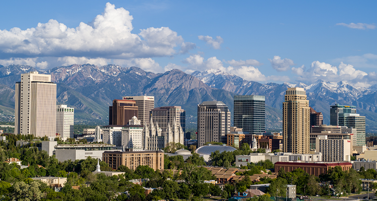 View of a city skyline with modern buildings and a prominent historic temple, set against mountains under a blue sky with clouds.