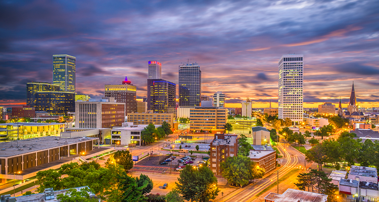 City skyline of Tulsa at sunset with tall buildings, colorful sky, and lit streets.