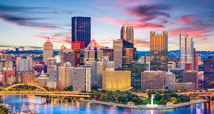 Pittsburgh skyline at sunset with illuminated skyscrapers, yellow bridges, and calm river reflecting city lights.