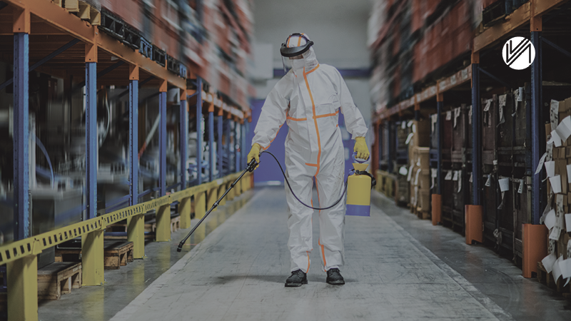 Person in white protective suit and gloves disinfecting a warehouse aisle with a sprayer.