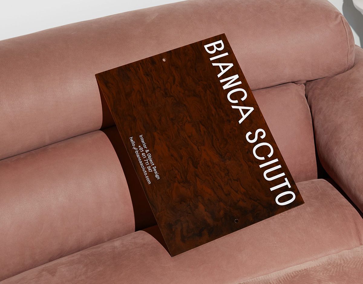 Dark woodgrain presentation folder with a vertical Bianca Sciuto wordmark, resting on a pink velvet sofa