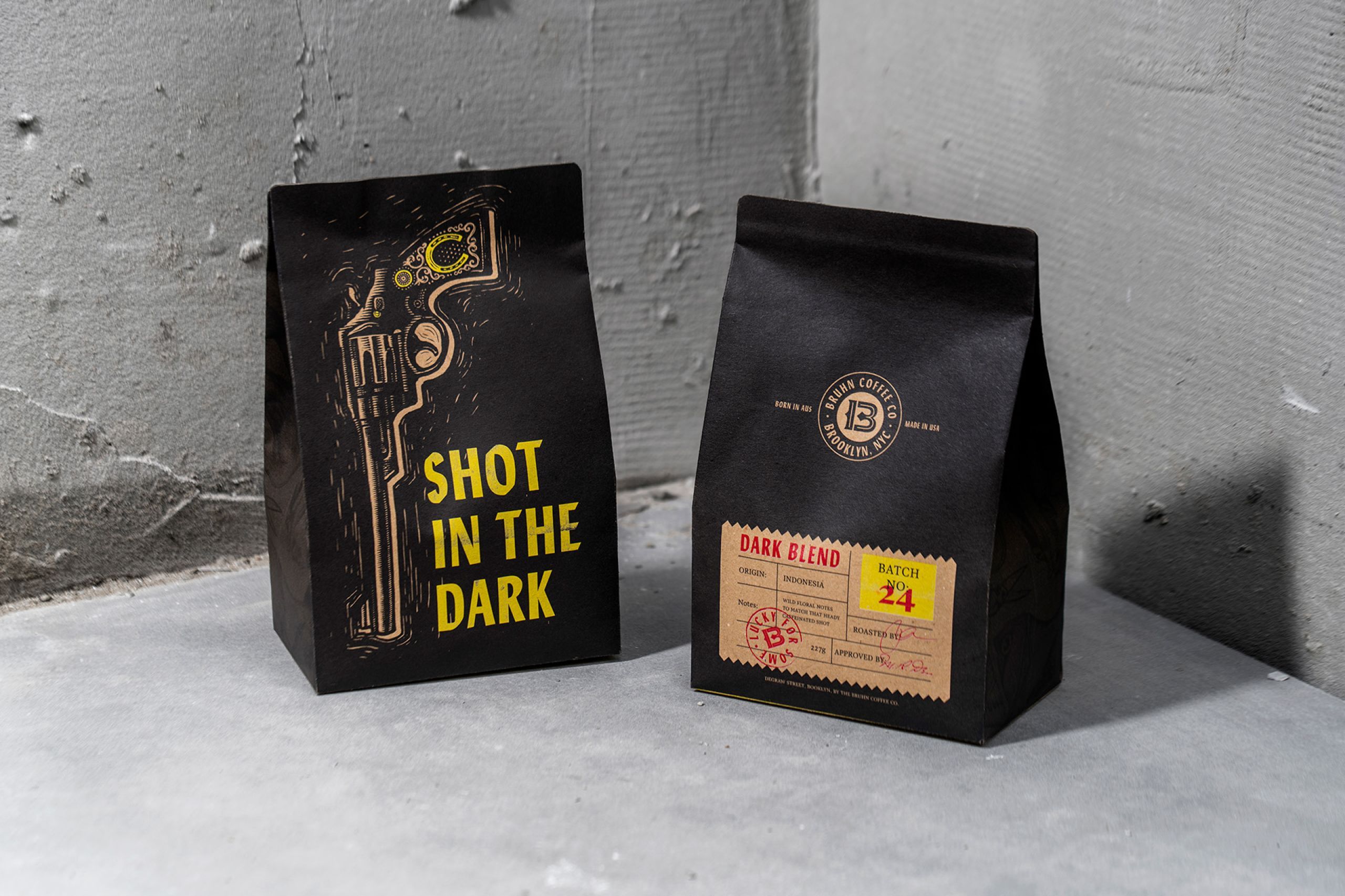 Two matte black coffee bags with yellow line illustration and a Shot in the Dark wordmark, photographed against a concrete wall