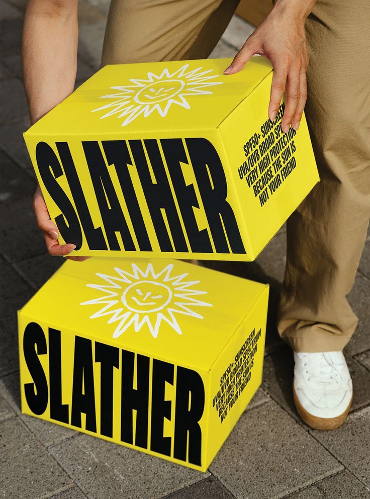 A person carrying two bright yellow Slather cartons printed with a sunburst motif and heavy black wordmark
