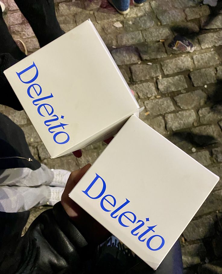 Two white boxes branded Deleito in blue serif type, held above wet cobblestones at night