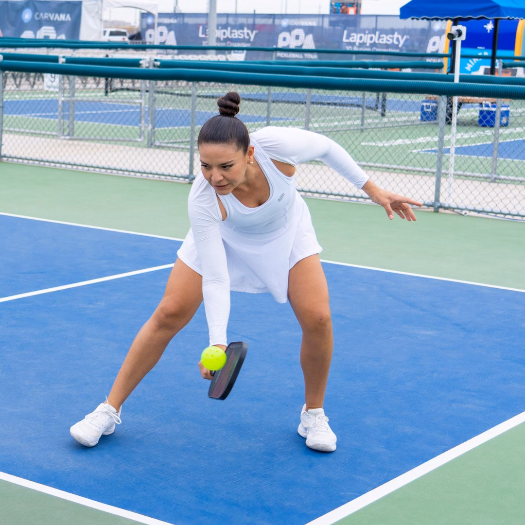 girl in white clothing hitting a one handed backhand dink on a blue pickleball court