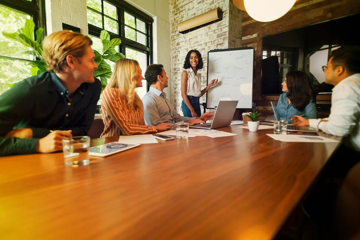 Mensen aan een tafel die samen overleggen en kijken naar een vrouw bij een whiteboard