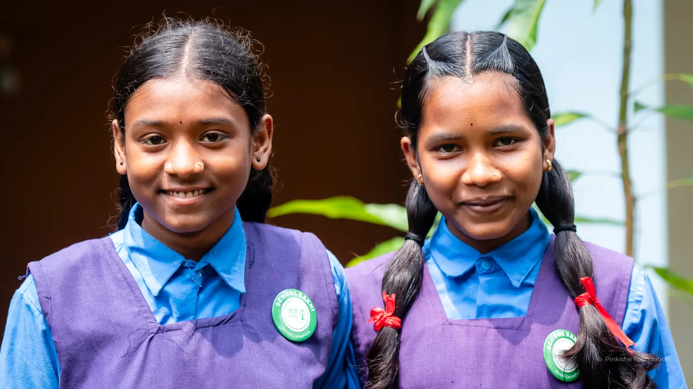 Two schoolgirls in uniform smiling outside their classroom at a Pinkishe supported school