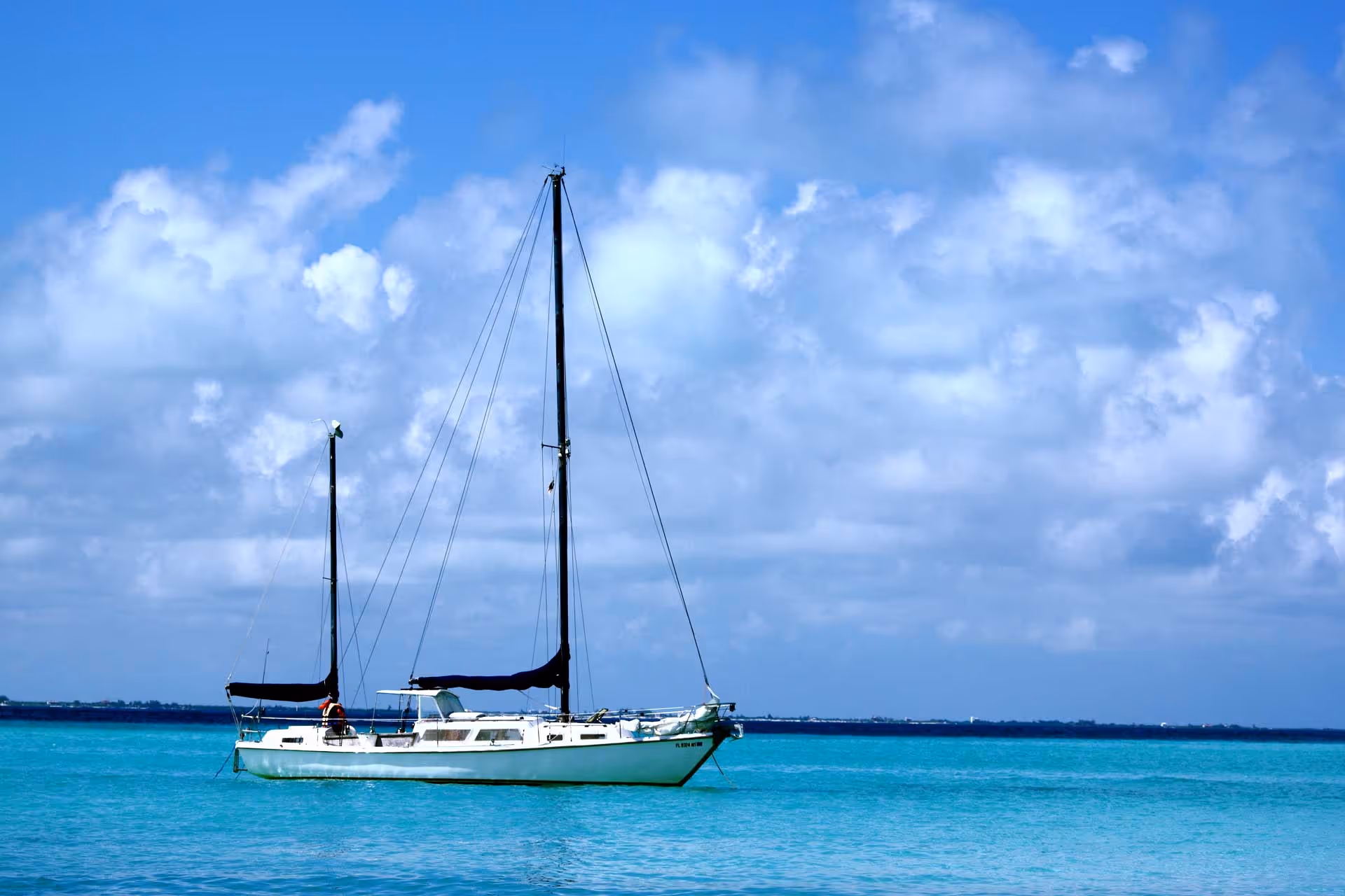White sailing yacht anchored in clear turquoise waters near a lush green island with rocky cliffs on a sunny day.