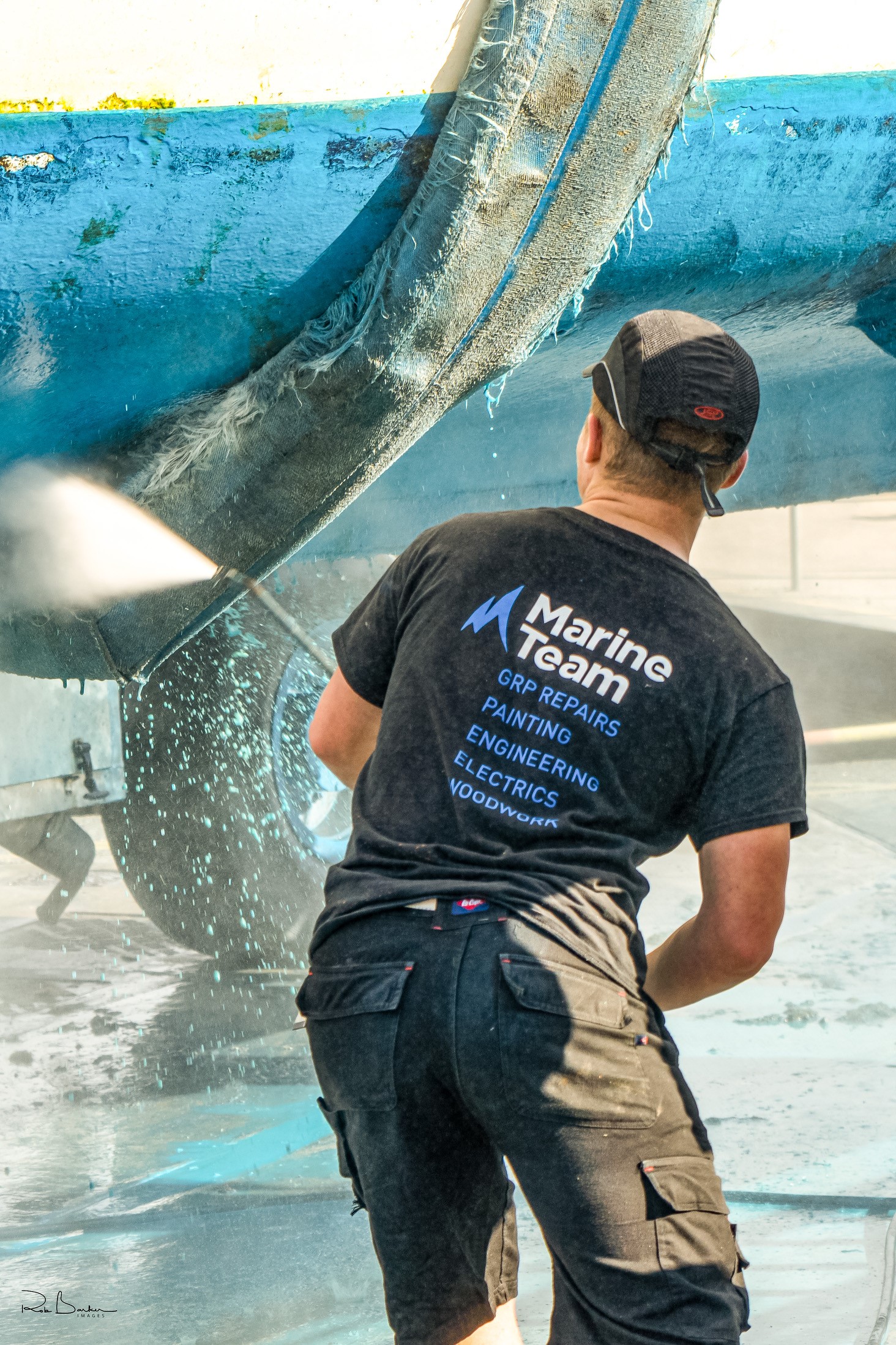 Marine technician pressure washing the underside of a sailboat hull and keel in a boatyard, wearing a Marine Team branded work shirt.