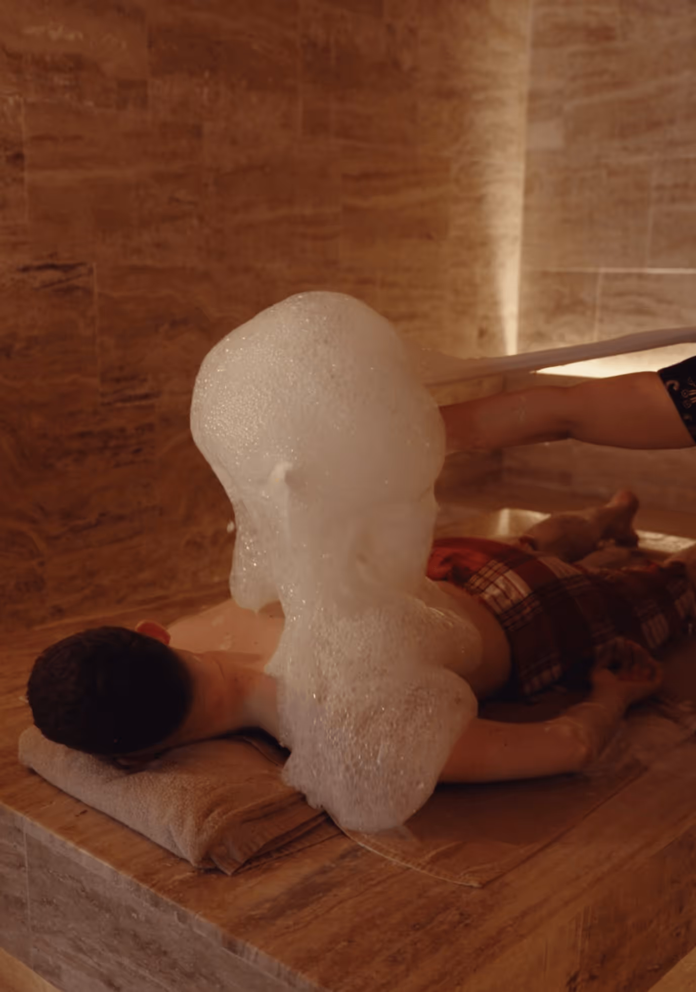 Person lying face down on a stone surface with a towel under their head while receiving a foam massage in a spa setting.
