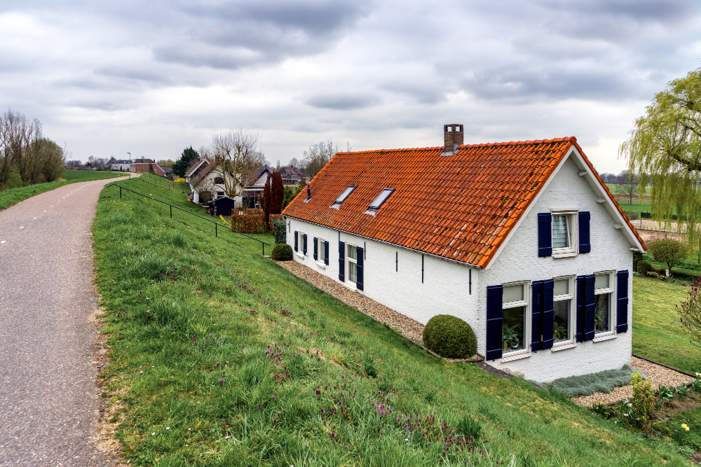 Une maison de campagne traditionnelle aux murs blancs et toit de tuiles terre cuite orange, située au pied d'un grand talus herbeux vert sous un ciel couvert. Un chemin mène vers d'autres habitations.