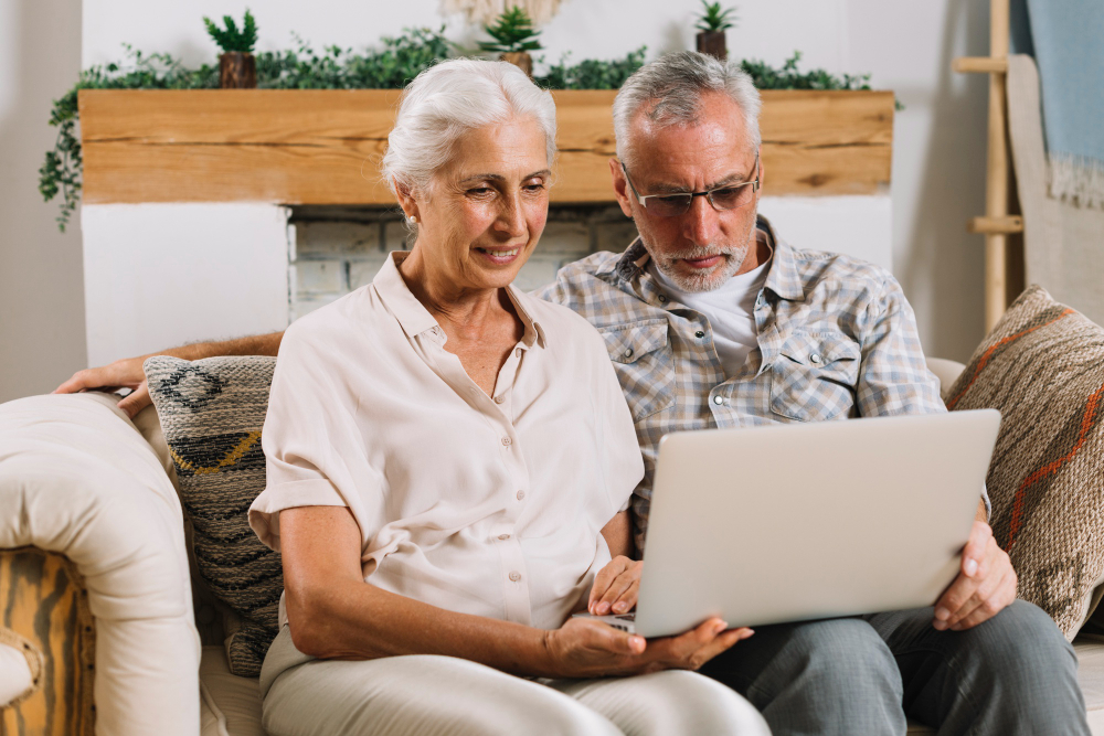 Un couple de retraités souriant sur son canapé, effectuant des recherches en ligne sur un ordinateur pour trouver une aide au logement senior.