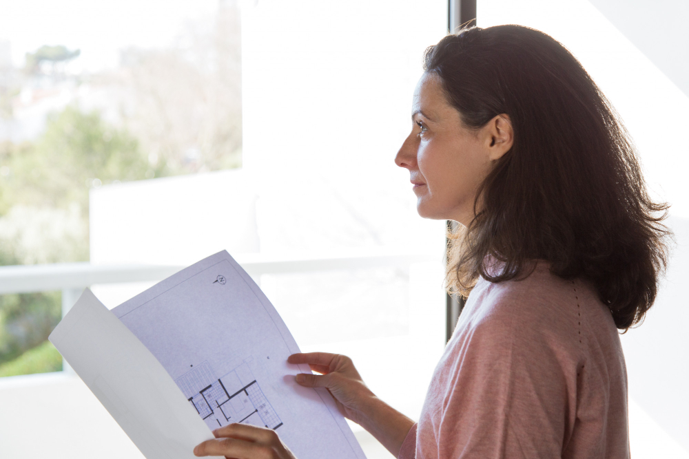 Profil d'une femme analysant des plans d'architecte devant une fenêtre lumineuse, dans le cadre d'un projet de construction RE2020.