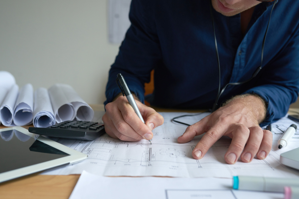 Gros plan des mains d'un architecte dessinant avec un stylo sur un plan technique, bureau avec calculatrice et tablette pour une étude thermique.