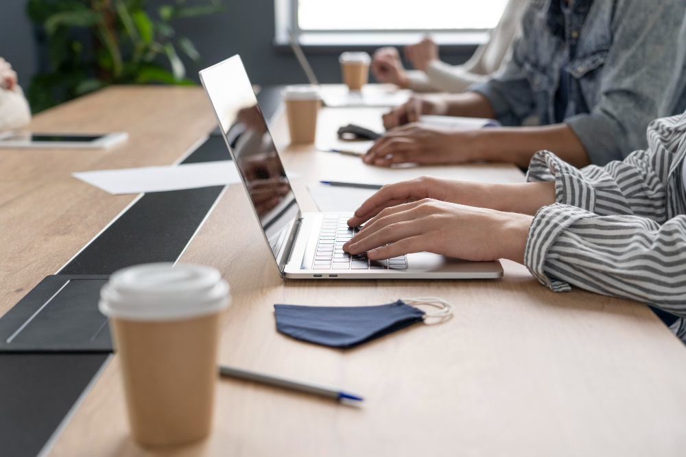 Gros plan sur un coworker tapant sur un clavier d'ordinateur portable dans un bureau partagé à Chartres-de-Bretagne, avec un gobelet de café et un masque sur la table.