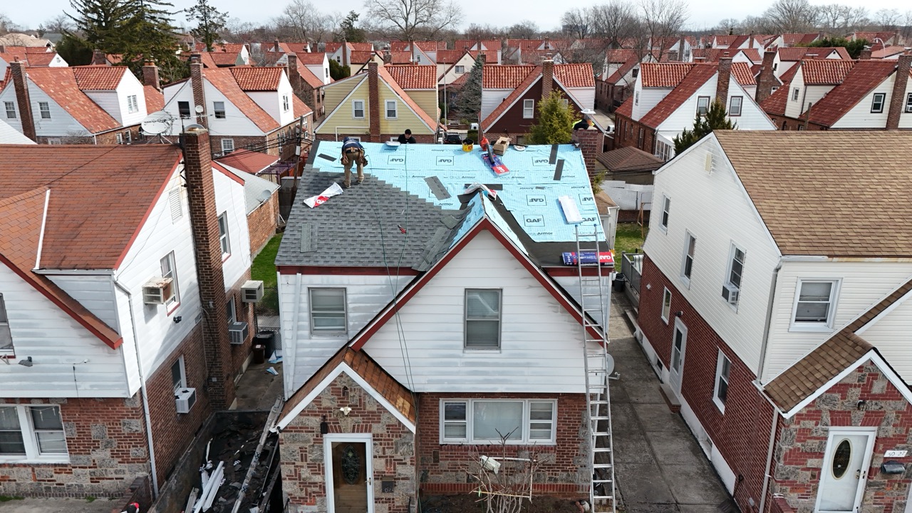 Workers installing grey shingles on a residential roof under construction in a neighborhood of brick and siding houses.