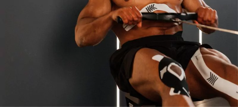Knie tapen Titelbild. Ein Mann sitzt an einem Rudergerät im Fitnessstudio. Man sieht sein getaptes Knie. Das Tape hilft ihm beim Stabilisieren des Knies.