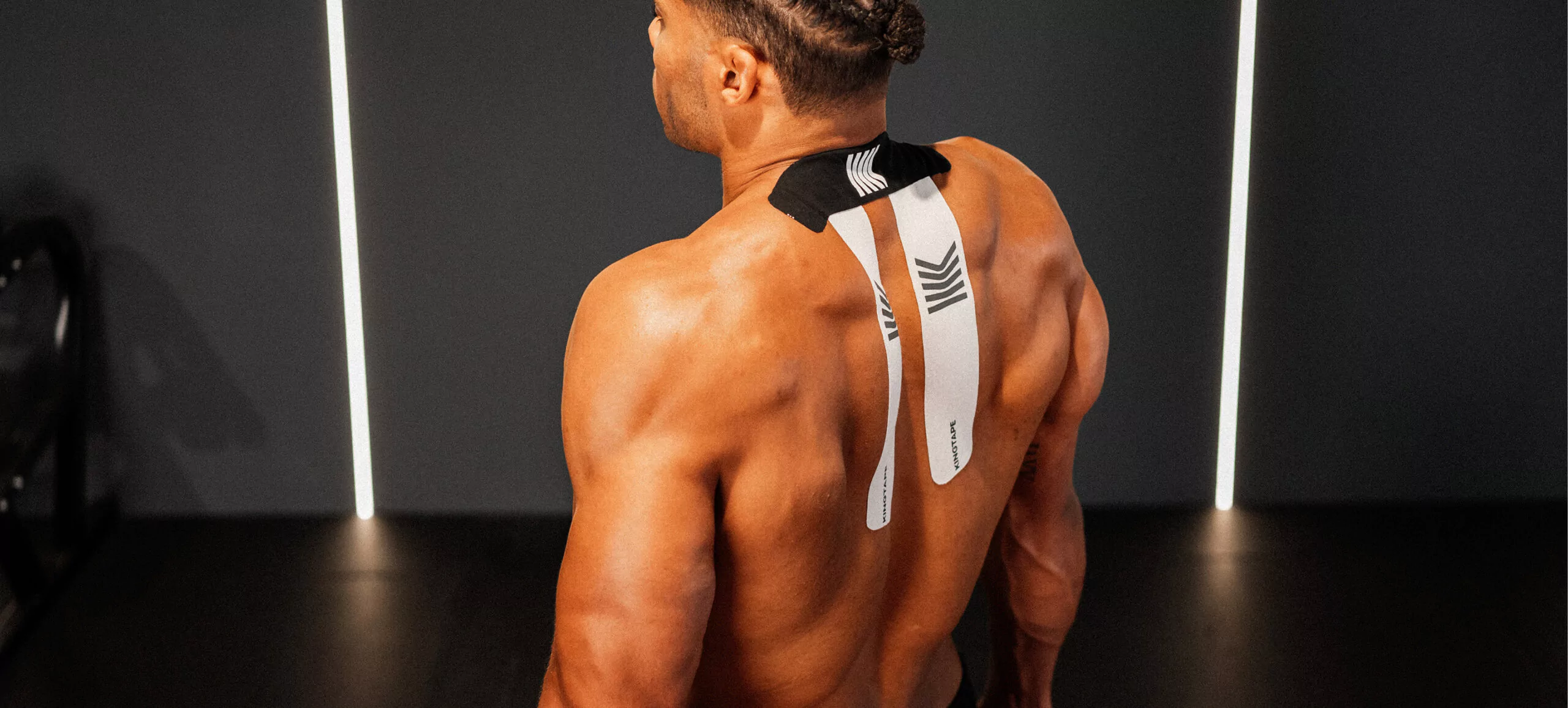 Mann im Fitnessstudio von hinten fotografiert, trägt ein Kinesiologie-Tape im Nacken- und Schulterbereich zur Linderung von Nackenverspannungen.