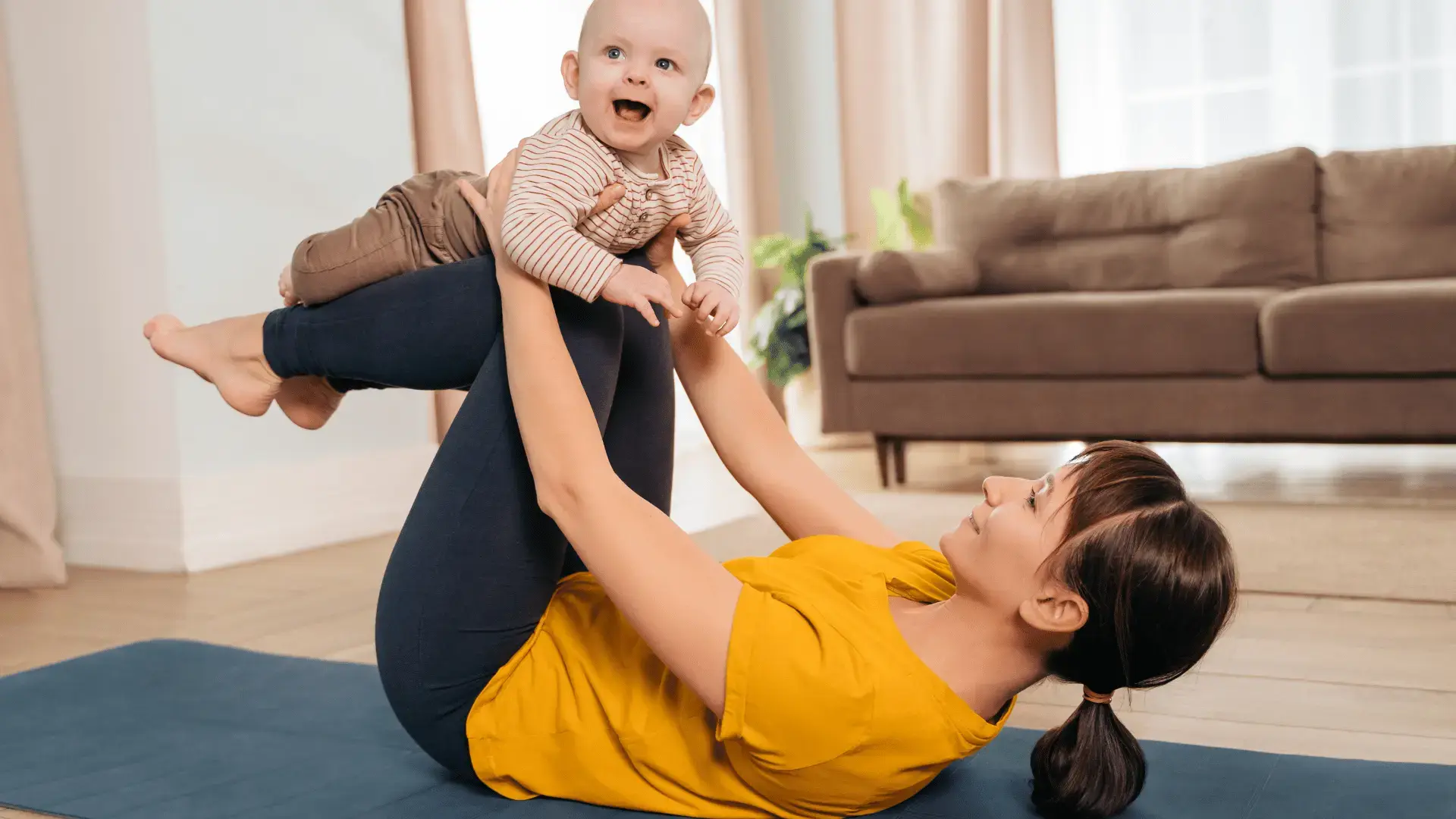 Eine Frau liegt auf einer Yogamatte im Wohnzimmer und hält ein lachendes Baby freudig in die Höhe