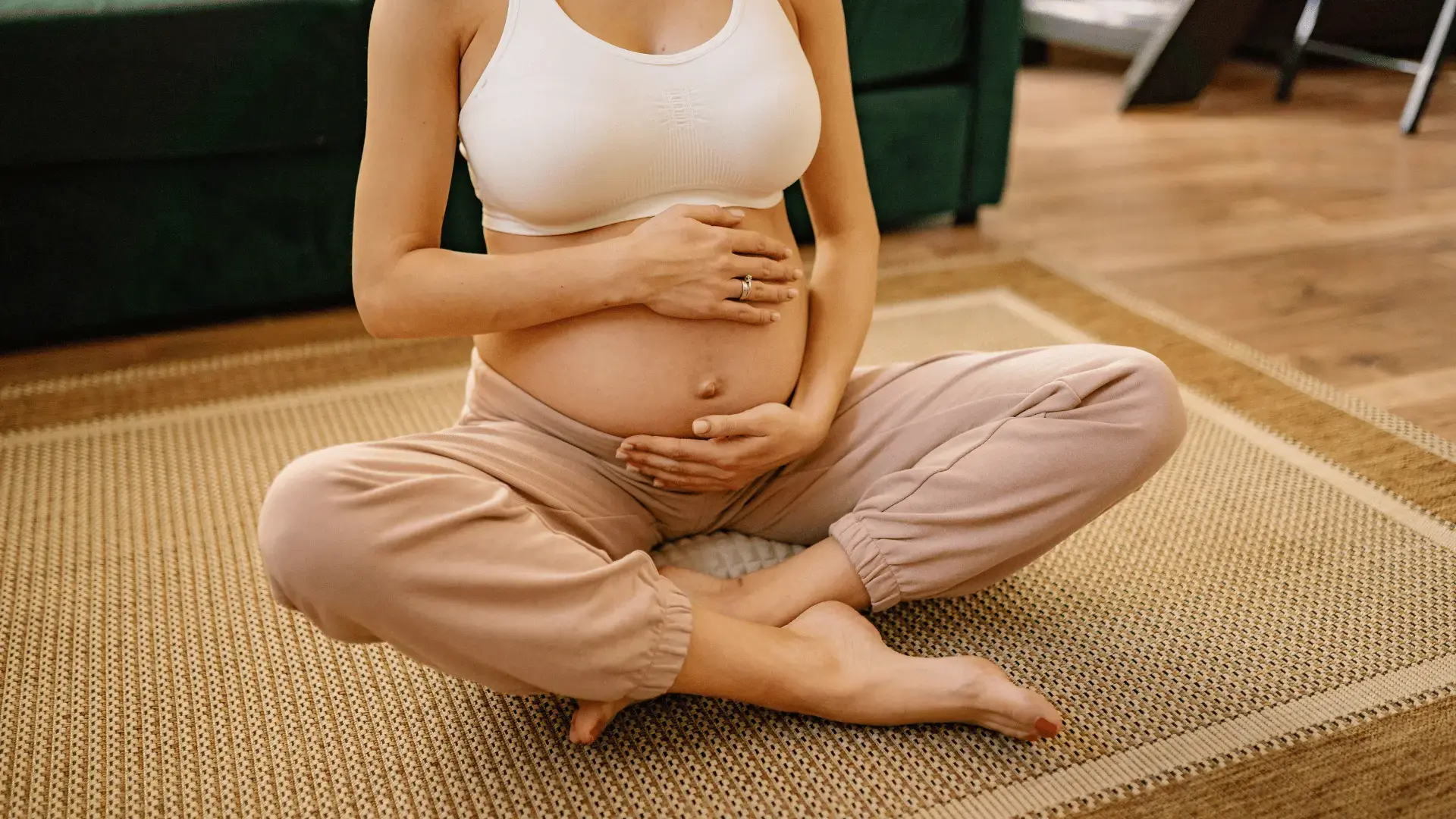 Hier ist ein kurzer, präziser und SEO-freundlicher Alt-Text für das Bild: Alt-Text: Schwangere, sportliche Frau sitzt entspannt im Schneidersitz auf einer Bodenmatte und umfasst behutsam ihren bloßen Babybauch.